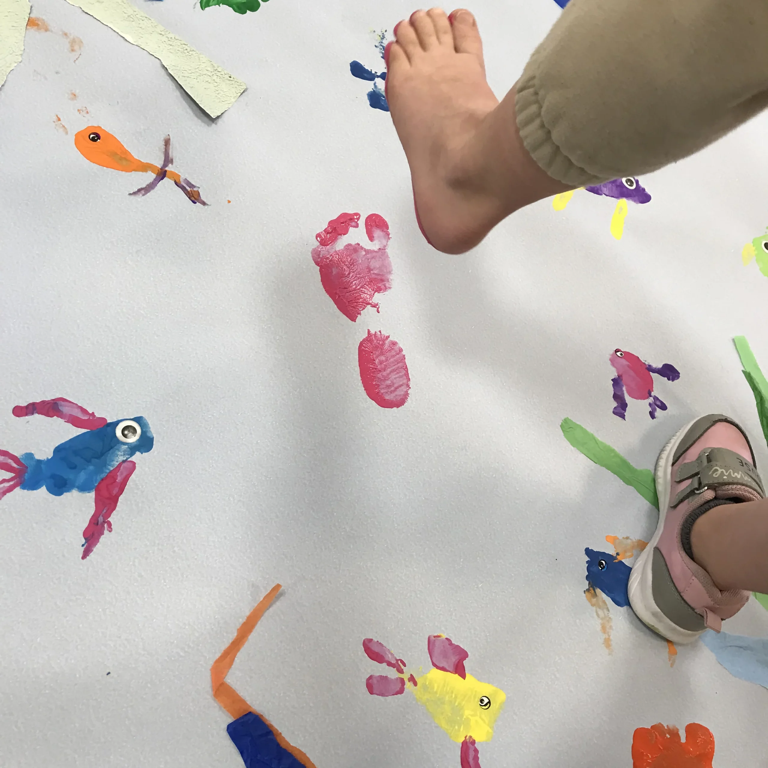 Projet de fresque rivière avec l'école maternelle Salengro à Orchies. On voit un pied d'enfant qui se soulève, laissant une empreinte de pas rouge sur le papier de la fresque. Autour d'autres formes de mains et de pieds ont déjà été décorées afin de ressembler à des poissons bariolés.