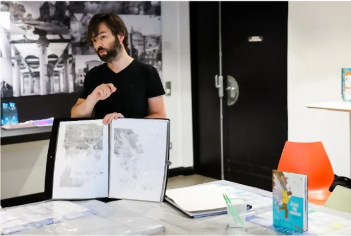 On peut voir Julien Martinière dans une salle de médiathèque en train d'expliquer les techniques qu'il utilise dans ses albums. Il tient ouvert un carnet de croquis où des crayonnés sont visibles. Sur la table devant lui, d'autres de ses dessins sont posés et un de ses albums est présent. Crédit photo : Kalimba.