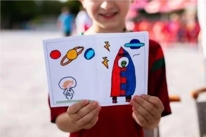 Un enfant avec un t-shirt rouge tient devant lui la page qu'il a réalisé de l'atelier Myrmidon. On y voit le personnage de Myrmidon en tenue de spacionaute, au dessus de lui il y a des planètes (rouge, jaune avec anneau et bleu) des éclairs et à sa droite, une fusée bleue et rouge dans laquelle se trouve un alien vert. Crédit photo : Kalimba