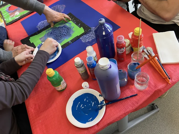 On voit les enfants utiliser des pochoirs avec de la peinture bleue claire sur fond foncé afin de réaliser le feuillage de leur tableau. Le matériel de peinture est en premier plan.