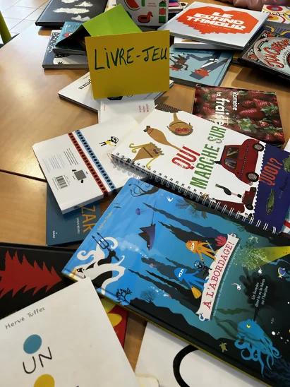 On voit une pile de livres-jeux sur la table, des méli-mélo, des livres dont tu es le héros, des cherche et trouve, etc. Une étiquette "livre-jeu" est posée sur la pile.