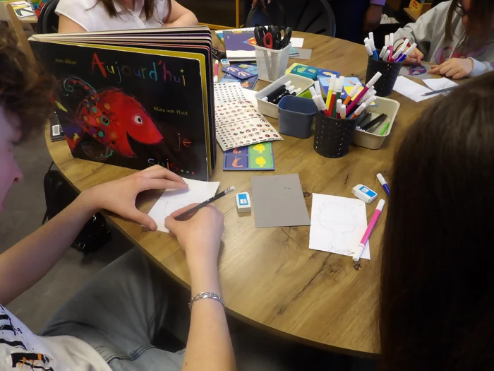 On voit des jeunes en train de créer leurs cartes poissons pour l'atelier, sur la table de la papeterie est à leur disposition pour dessiner. Le livre "aujourd'hui je suis" est sur la table, il y a un gros poisson multicolore mais principalement rouge en couverture.