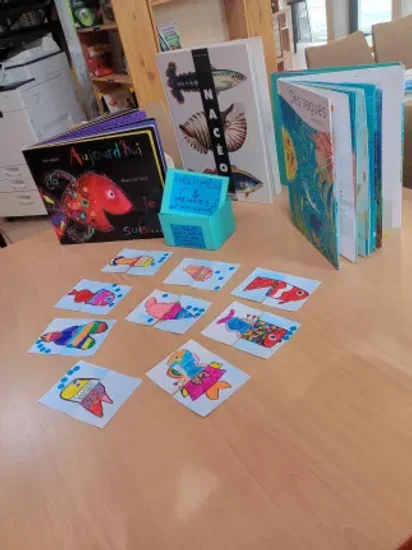 On voit un set de cartes de poissons dont les moitiés ont été mélangées. La boite du jeu ( bleue) est en enxposition derrière, avec les livres en lien avec l'atelier ( aujourdh'ui je suis... , Nacéo et Des vagues).