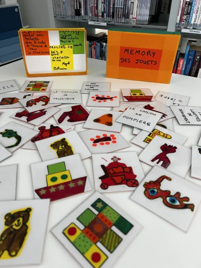 On voit les cartes du memory jeux et jouets finies. Des nounours, des bateaux de bain, des lunettes rigolotes, des quilles, des cubes, etc sont représentés sur les cartes. Derrière la boîte de rangement du jeu est en exposition. Elle est aux couleurs de Perluette.