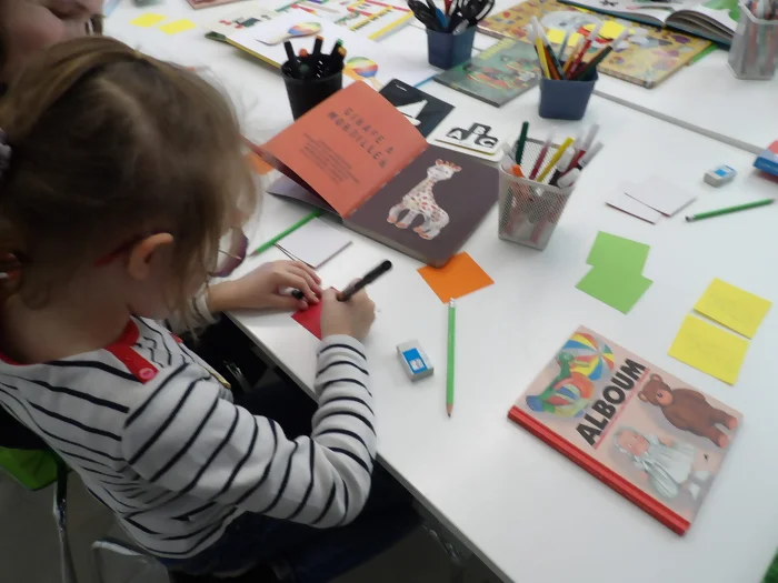 Une enfant est en train de dessiner sa carte de memory en se basant sur une illustrations de Sophie la giraffe dans un livre.