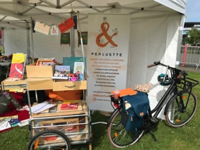 On y voit une tente blanche, sous laquelle une banderole Perluette est en fond. Au premier plan, une pile de livre dans une étagère à roulettes tirée par un vélo. C'est le projet de bibliothèque nomade proposé ar Perluette