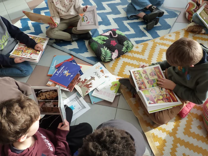 On voit un groupe d'enfants, assis sur des tapis de lecture colorés. Ils sont en train de lire des bandes dessinées muettes (qui n'ont pas de bulle de texte) provenant de la malle créée par Perluette