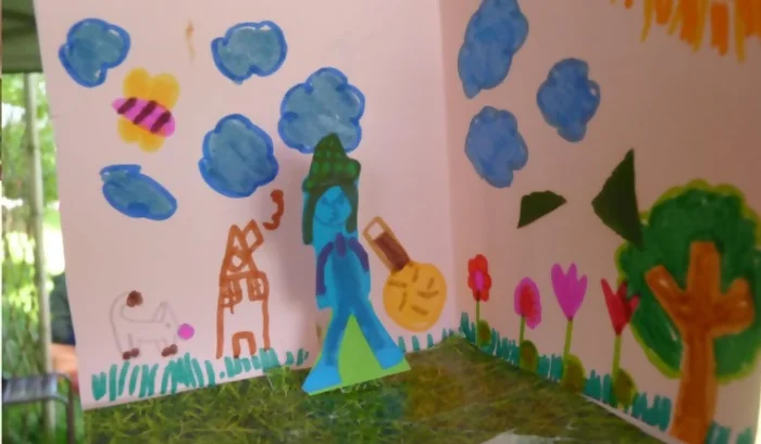 Exemple de carte pop-up à croquer : on voit que le fond a été dessinée aux feutres, des nuages bleus et des insectes volent dans le ciel. Un soleil brille au dessus d'un jardin avec un arbre fruitier et des fleurs. Une petite maison est dessinée sur la gauche avec un animal. Le sol est fait avec une image d'herbe découpée et collée sur le fond de la carte. Un personnage en train de jardiner apparaît en relief, tenant une tondeuse à la main.