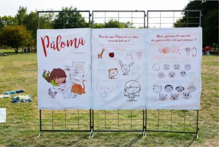 Panneaux de l'exposition Paloma, placés dans un jardin sur des grilles. On voit le titre "Paloma" en rouge avec les personnages dessinés en dessous en train de faire les ateliers de l'exposition.