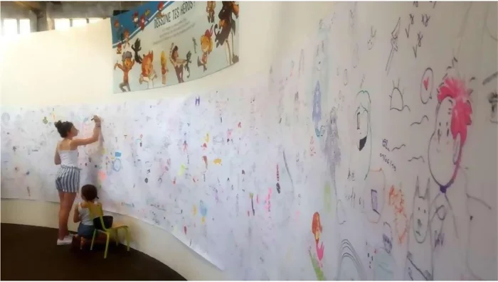 Une grande fresque sur un mur arrondi recouvert de dessin des visiteurs sur les héros et héroïnes. Au fond une femme et une petite fille ajoutent leur dessin. Des crayons posés sur une petite chaise sont à disposition pour dessiner. Crédit photo : Kalimba