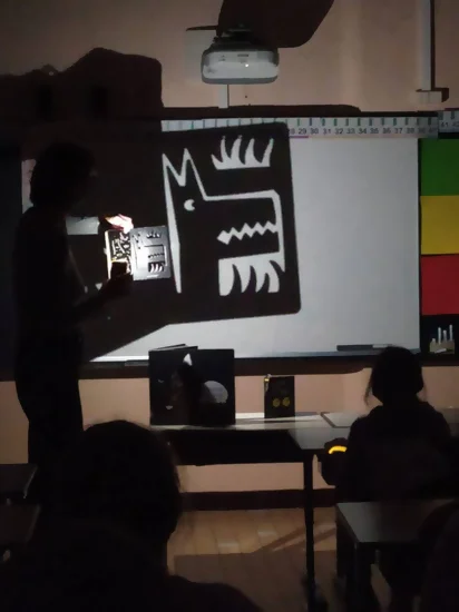 On voit Laure Montaigne en train de projeter une page d'album en papier dentelle à l'aide d'une lampe de poche sur un tableau blanc dans une salle de classe plonger dans le noir. Les enfants écoutent l'histoire en suivant avec les ombres des dessins projetés. Cet atelier a pris place dans le cadre des nuits de la lecture.