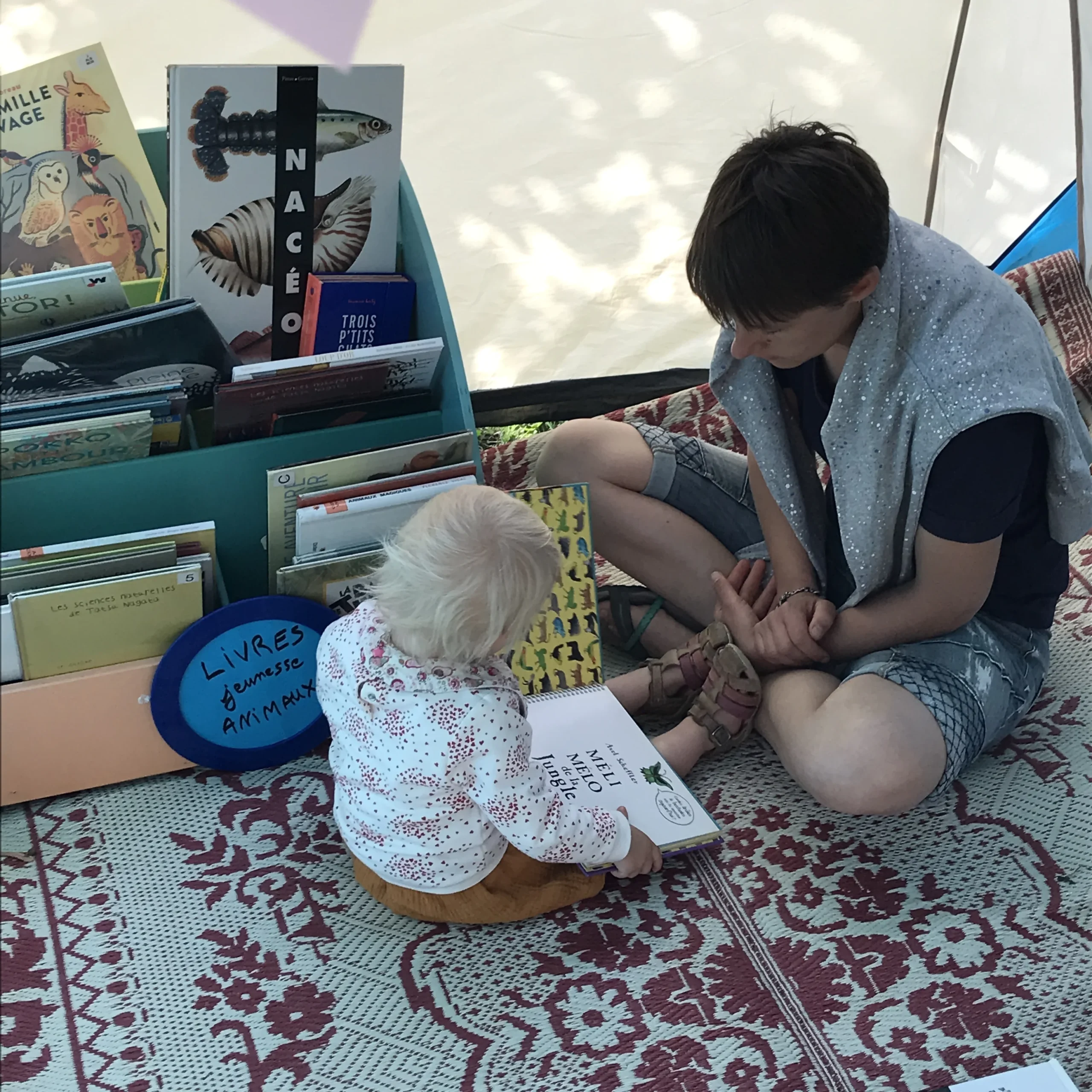 On voit un parent avec un enfant en bas âge en train de lire un album méli-mélo assis sur un tapis de lecture. Ils se trouvent dans une tente dressée par Perluette pour le festival Partir en Livre 2021