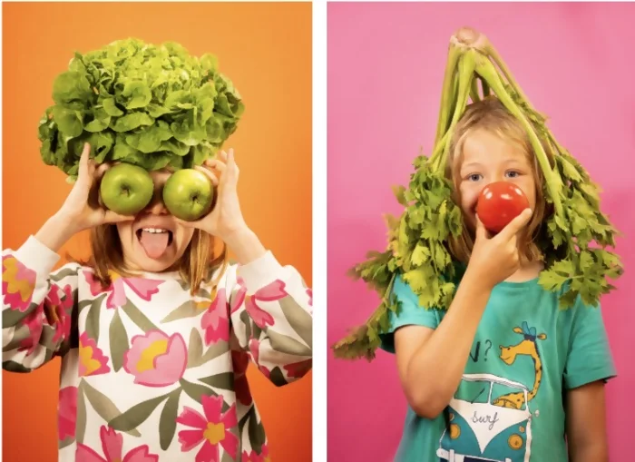 Portraits de deux enfants faits pendant l'atelier. Une des petite fille tient deux pommes vertes devant ses yeux, et à une salade de laitue en guise de cheveux. Elle tire la langue et porte un t-shirt blanc avec des fleurs roses aux feuilles grises. Elle se trouve sur un fond orange. La deuxième enfant a un céleri retourné sur la tête comme chapeau et tient une tomate rouge sur son nez, comme un clown. Elle porte un t-shirt bleu avec une girafe dessus. Elle se trouve sur un fond rose. Crédit photo : Kalimba