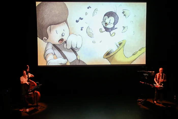 Perluette - spectacle Billy Symphony - vue de la scène On voit la scène du spectacle, une image de la bande dessinée est projetée sur l'écran, Billy vient de rouver un petit oiseau dans son saxophone, sortant juste de sa coquille. Sur la scène autour de l'écran, on voit la contrebassiste et le saxophoniste jouer devant le public dans une lumière tamisée