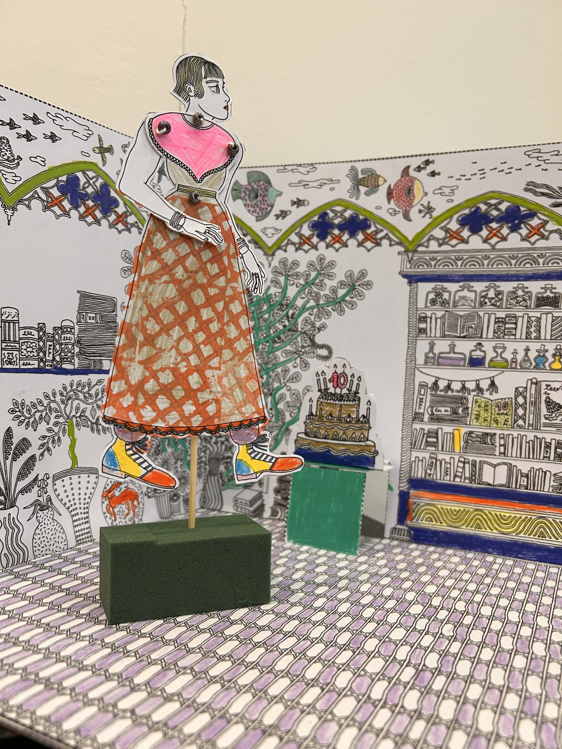 Carte pop-up créée par l'illustratrice Adley à l'occasion des 10 ans des Nuits des Bibliothèques de la MEL. Un personnage de lectrice est disposée debout dans un décor de bibliothèque "empoissonnée". Un gâteau d'anniversaire apparaît au second plan.