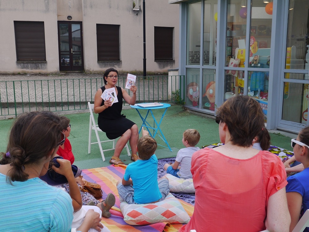 Mornant1_24juin17 Une comédienne présente trois romans de la collection "J'aime lire" des éditions Bayard devant des enfants assis sur un tapis avec des coussins et des adultes derrière sur des chaises. En plein air, dans la cour d'une librairie.