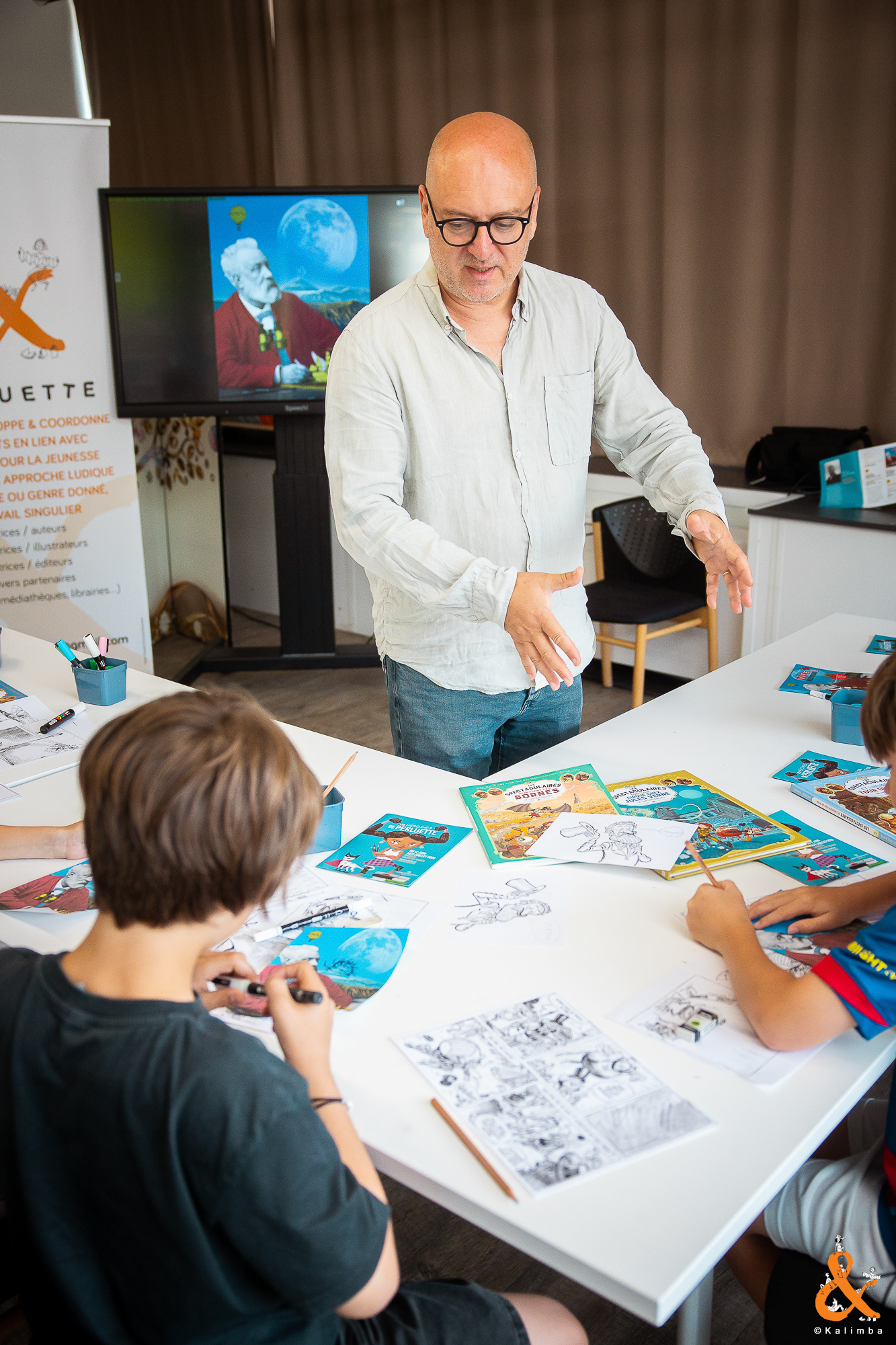Régis Huatière en train d'expliquer à deux enfants les codes de la bande dessinée. Les enfants tentent de reproduirent sur leurs exemplaires ces codes. Des flyers "Les Héro.ïne.s de Perluette" et les livres des "pestaculaires" de Régis Hautière sont autour d'eux sur la table.