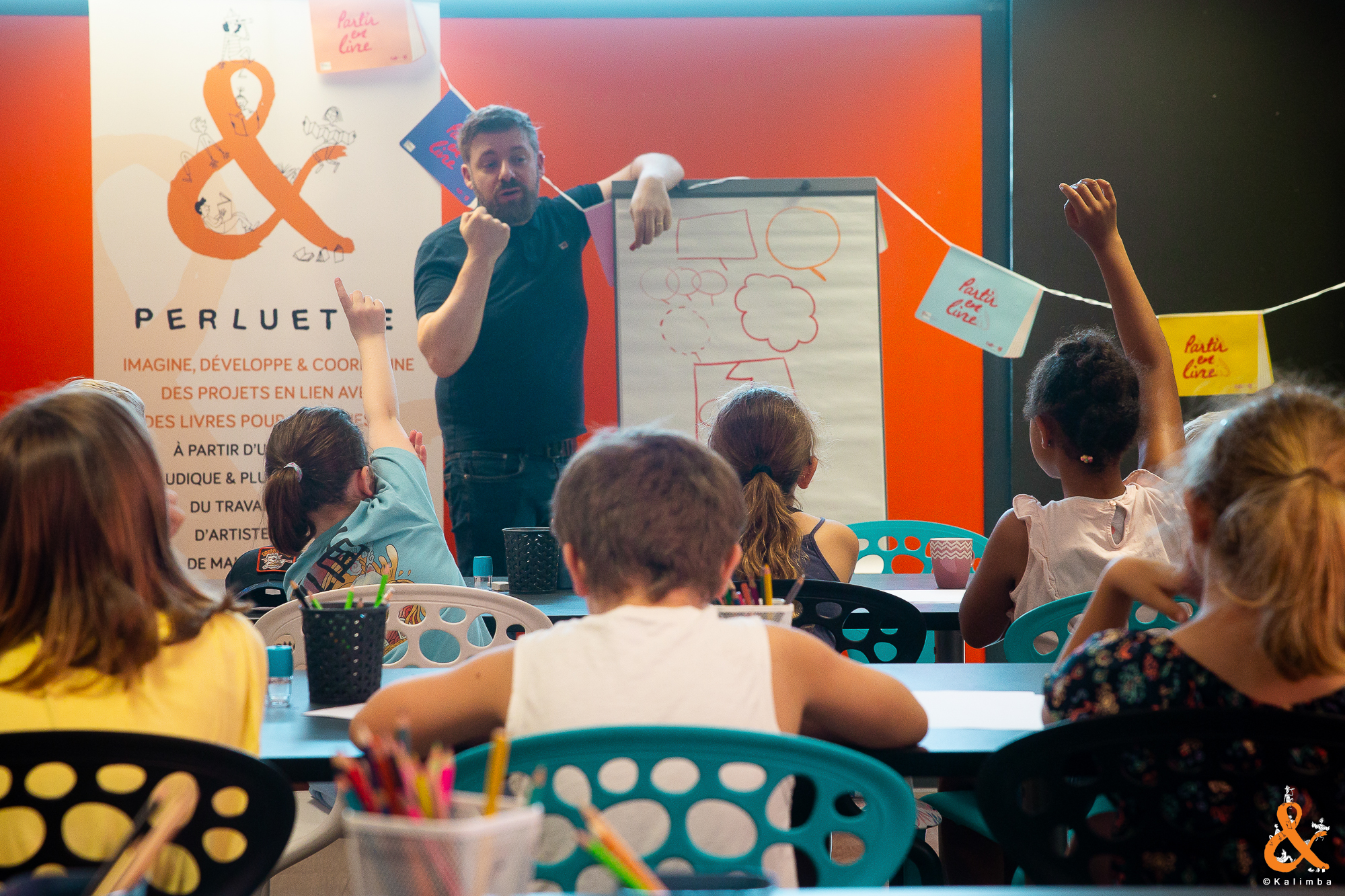 Le dessinateur Thomas Priou explique l'utilisation des différentes bulles de dialogue des bandes dessinées. Un calico Perluette et des banderolles Partir en Livre entourent le paperboard sur lequel il dessine. En premier lan on voit des enfants assis à des tables qui l'écoutent et lèvent la main pour poser des questions. crédit photo Kalimba