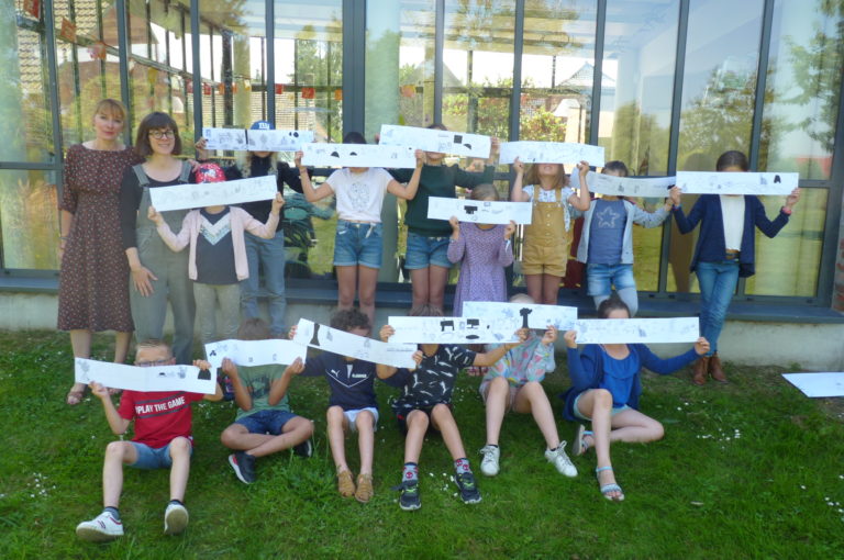 Les enfants tiennent les livres accordéons qu'ils ont réalisés devant leur visage. Magali Dulain est à leur droite avec la médiathécaire. La photo prend place dehors sur l'herbe.