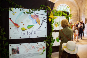 Entrée de l'exposition "la toile" issue du travail d'Isabelle Simler dans l'album du même nom. On voit un panneau avec le titre "la toile" marqué en bas en noir dans une calligraphie un peu patte-de-mouche. Au dessus du titre, des insectes sont dessinés : papillon, puceron, coccinelle, libellule. Le panneau est posé sur une grille couverte de tissus noirs et de faux lierres pour donner un effet de nature. On voit d'autres panneaux similaires au fond de l'image avec des visiteurs en train de les lire. L'exposition semble se trouver dans un bâtiment fait de vieilles pierres.