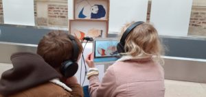 Deux enfants, une fille et un garçon sont assis devant l'exposition interactive, avec des casques audio sur les oreilles ils écoutent l'animation qui accompagne les dessins de l'autrice.