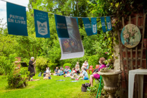 Nous sommes dans un jardin, une banderole Partir en Livre est suspendue à la haie en premier plan. Derrière, le public est assis dans l'herbe et sur des chaises de jardin à écouter une conteuse